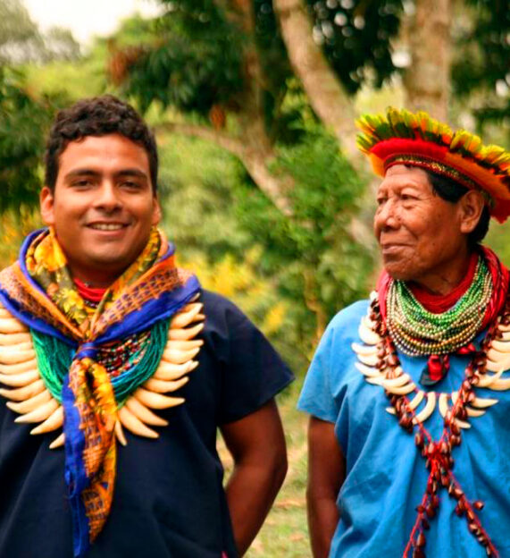 Two Indigenous men smiling, wearing traditional beaded necklaces with large curved claws or teeth, in front of a blurred jungle background.