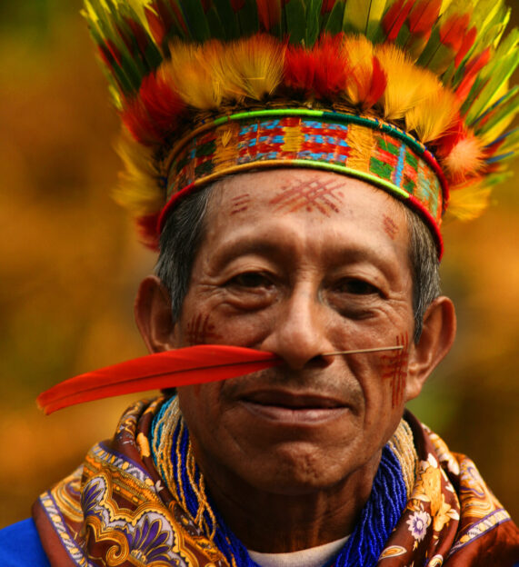 Headshot of an Indigenous man, smiling and wearing traditional feathered headgear with a red feather pierced through his nose.