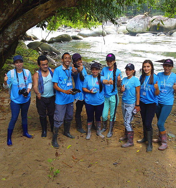 Savimbo Villagarzon team members, wearing matching turquoise shirts, stand under a large tree in front of a river in Putumayo Amazon, Colombia.