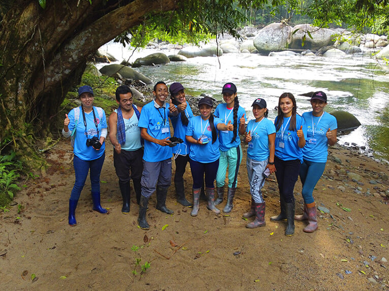 Savimbo Villagarzon team members, wearing matching turquoise shirts, stand under a large tree in front of a river in Putumayo Amazon, Colombia.