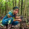 An Indigenous woman kneeling at the base of a thin tree that holds a camera trap in the Putumayo Amazon, Colombia.