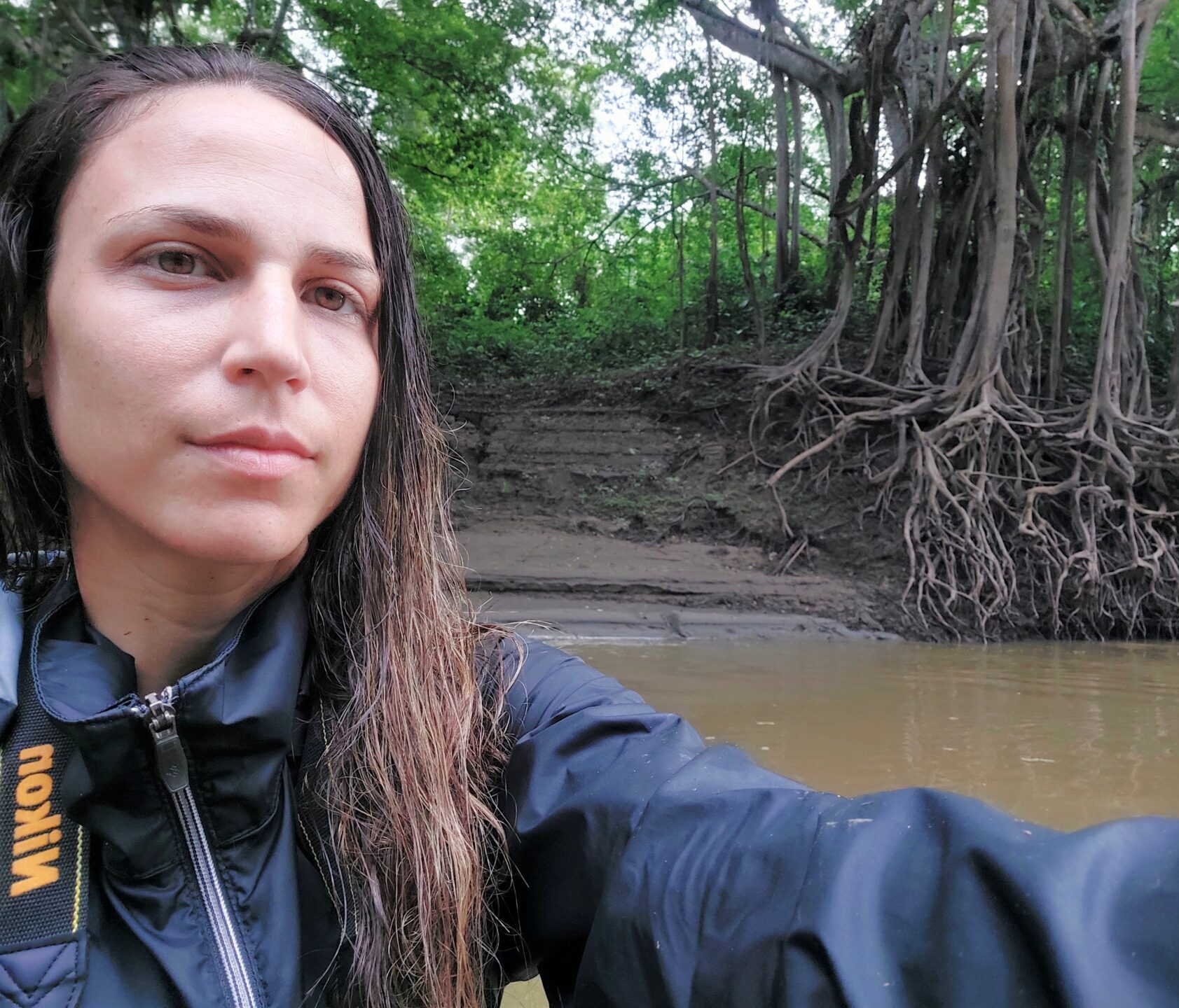Woman with long brown hair, wearing a dark blue raincoat and a camera around her neck, in front of a muddy river with trees in the background