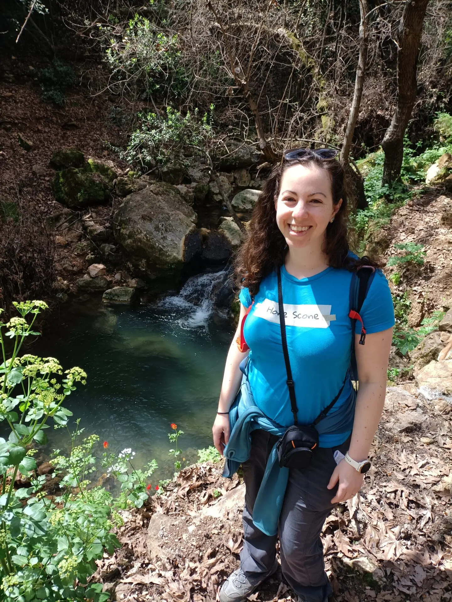 A smiling woman, standing beside flowering plants and a small river or pond.