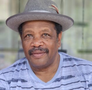 Portrait of an African man with a mustache, wearing a grey hat and a light blue striped shirt.
