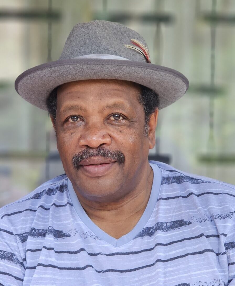 Portrait of an African man with a mustache, wearing a grey hat and a light blue striped shirt.
