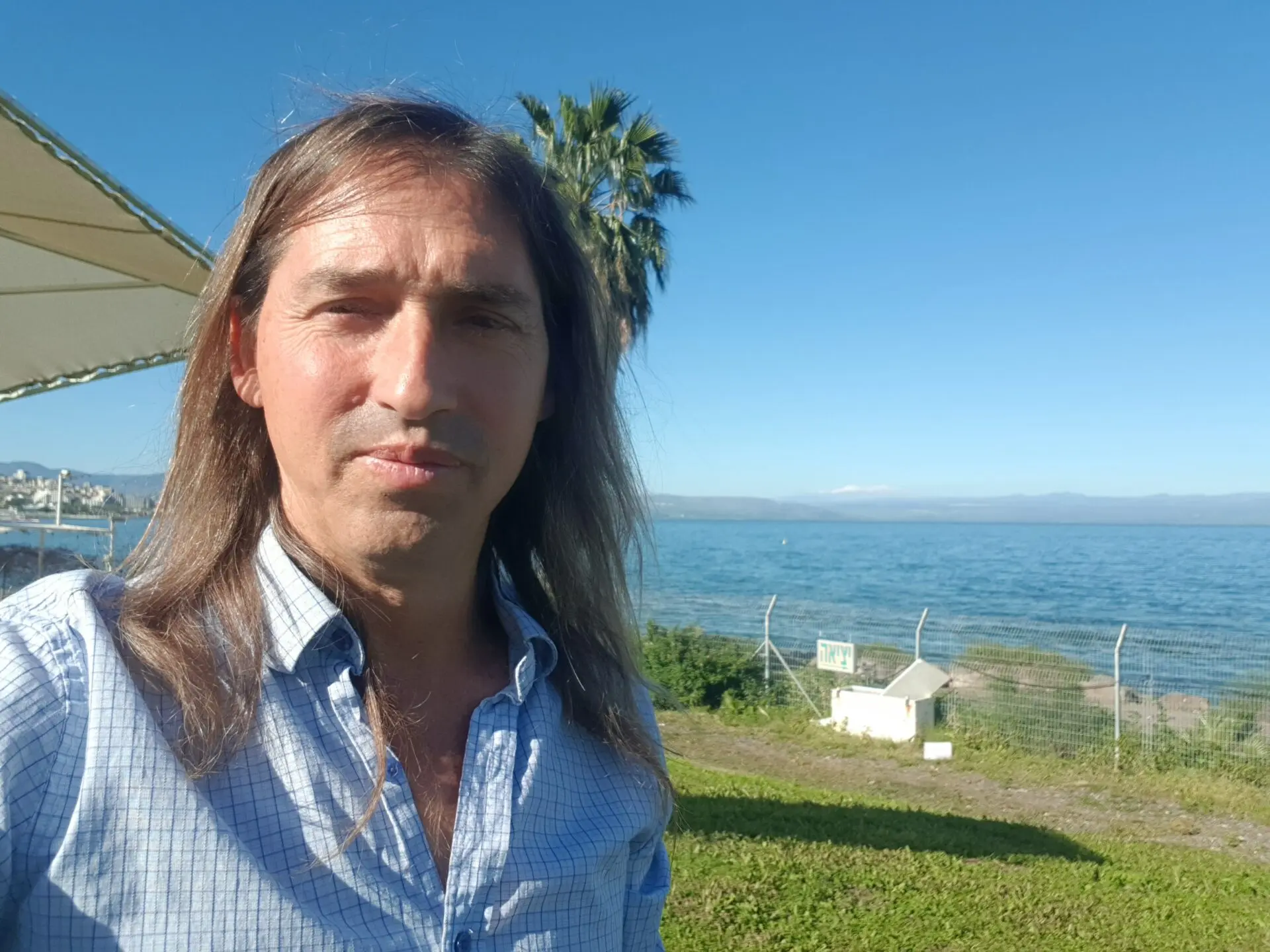 Close up photo of man with long brown hair with a field, a white house, and a body of water in the background