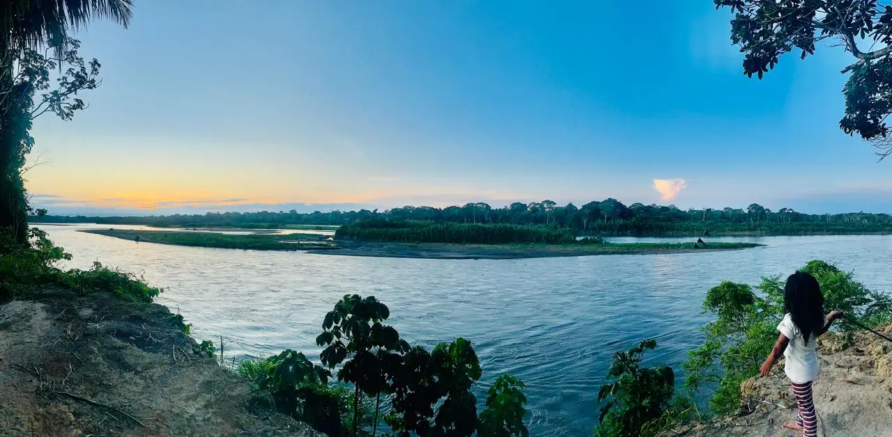 Panoramic photo of IKIAM Reserve, Ecuador, from the banks of a river, with a young girl looking out onto the river, a small island, and the sunset on the horizon.